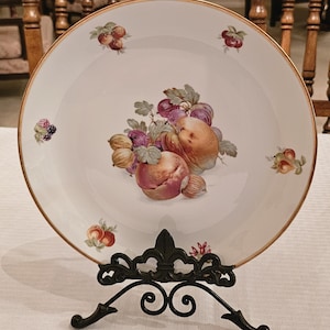 Könnte beinhalten: Ein weißer Porzellanteller mit Goldrand, verziert mit einem handgemalten Stillleben aus Pfirsichen, Trauben und anderen Früchten. Der Teller wird auf einem schwarzen Metallständer mit Fleur-de-Lis-Design präsentiert.
