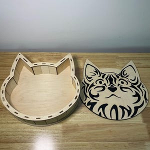 May include: A wooden cat-shaped box and lid. The box is light brown, with a detailed cat face design on the lid. The box is open, revealing the interior. The lid is separate, showing the cat's face in black and light brown.