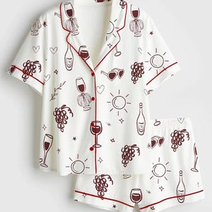 May include: White pajama set with a wine-themed pattern. The short-sleeved top and shorts feature a repeating design of wine glasses, bottles, grapes, and suns in a dark red color. The edges of the set are trimmed with a matching dark red piping.