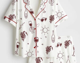 Op de afbeelding: Witte pyjamaset met een wijnthema. De top met korte mouwen en de shorts hebben een herhaald patroon van wijnglazen, flessen, druiven en harten in een bordeauxrode kleur. De set heeft bordeauxrode biezen.