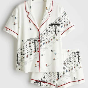 May include: White pajama set with a ski lift and skier print. The short-sleeved top and shorts have red trim and buttons. The illustration features skiers on a ski lift and on the slopes.
