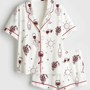 May include: White pajama set with a wine-themed pattern. The short-sleeved top and shorts feature a repeating design of wine glasses, bottles, grapes, and suns in a dark red color. The edges of the set are trimmed with a matching dark red piping.
