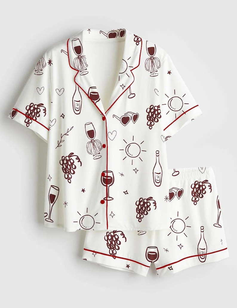 May include: White pajama set with a wine-themed pattern. The short-sleeved top and shorts feature a repeating design of wine glasses, bottles, grapes, and suns in a dark red color. The edges of the set are trimmed with a matching dark red piping.