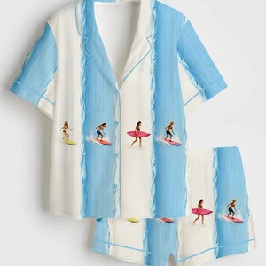 May include: A two-piece pajama set featuring a short-sleeved button-down shirt and matching shorts. The set has a white background with vertical blue stripes and illustrations of surfers riding waves. The edges are trimmed with a light blue piping.