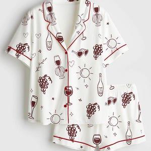 May include: White pajama set with a wine theme. The short-sleeved top and shorts feature a repeating pattern of wine glasses, bottles, grapes, and hearts, outlined in dark red. The edges of the set are trimmed in dark red.