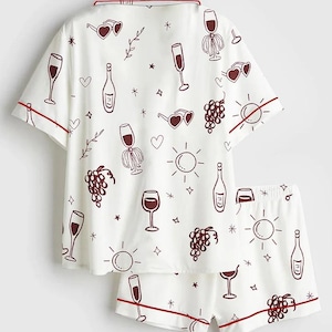 May include: White pajama set with a wine-themed pattern. The short-sleeved top and shorts feature a repeating design of wine glasses, bottles, grapes, and sunglasses. Red trim accents the edges of the set.