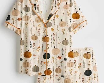 Könnte beinhalten: Cremefarbenes Pyjama-Set mit Kürbis- und Blumenmuster. Das kurzärmelige Oberteil und die Shorts haben einen orangefarbenen Besatz. Das Design umfasst verschiedene Kürbisse in Braun-, Orange- und Grüntönen sowie kleine Blumen und Blätter.