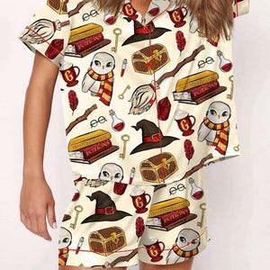 May include: Cream-colored pajama set with a repeating pattern of wizarding-themed illustrations. The design includes owls, books, keys, potion bottles, and witch hats. The set has red trim and a button-down shirt with matching shorts.