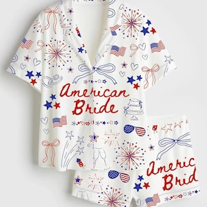 May include: White pajama set with a short-sleeved button-down shirt and matching shorts. The set is decorated with red, white, and blue illustrations of fireworks, stars, bows, and American flags. The words "American Bride" are written in red.