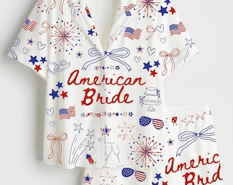 May include: White pajama set with a short-sleeved button-down shirt and matching shorts. The set is decorated with red, white, and blue illustrations of fireworks, stars, bows, and American flags. The words "American Bride" are written in red.