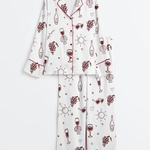 May include: White pajama set with a burgundy wine-themed pattern. The long-sleeved top and pants feature illustrations of wine bottles, glasses, grapes, and hearts. Burgundy trim accents the collar, cuffs, and pant hems.