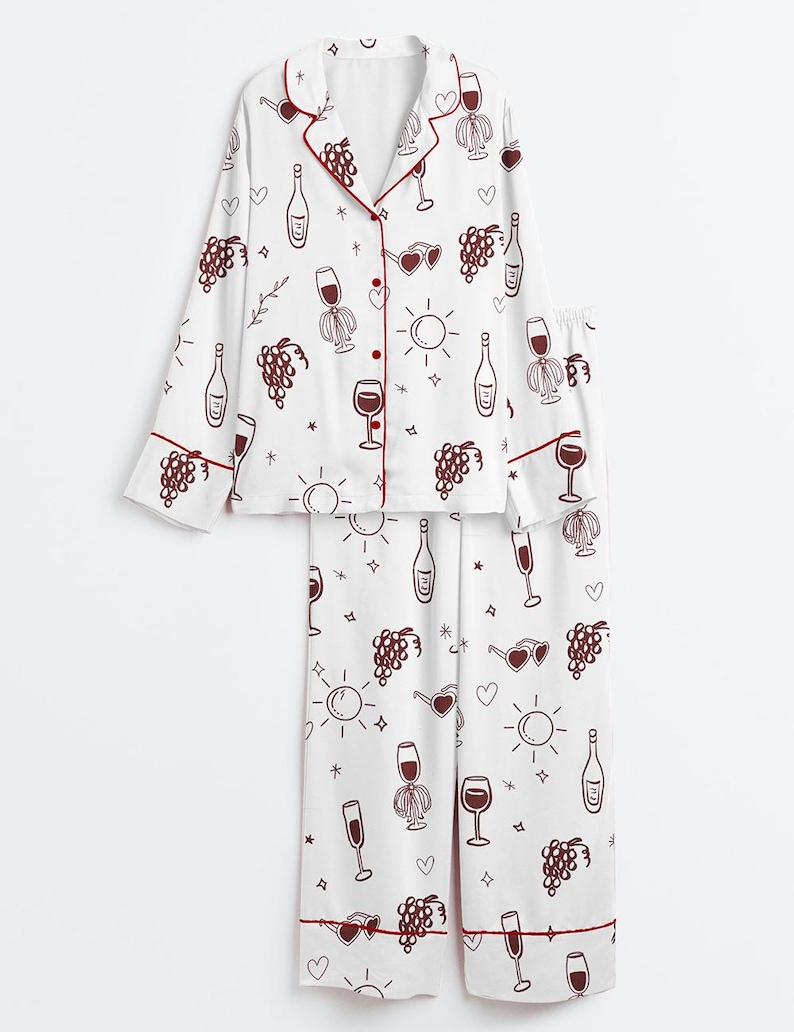 May include: White pajama set with a burgundy wine-themed pattern. The long-sleeved top and pants feature illustrations of wine bottles, glasses, grapes, and hearts. Burgundy trim accents the collar, cuffs, and pant hems.
