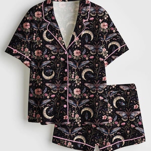 May include: Black pajama set with a floral, moth, and crescent moon pattern. The short-sleeved top and shorts are trimmed with pink piping. The fabric features a dark background with pink, blue, and white accents.