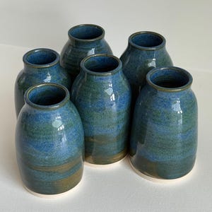 May include: Six handmade ceramic vases in a rounded shape. The vases are glazed in shades of blue and green, with a glossy finish. The base of each vase is a light cream colour. The vases are arranged together.