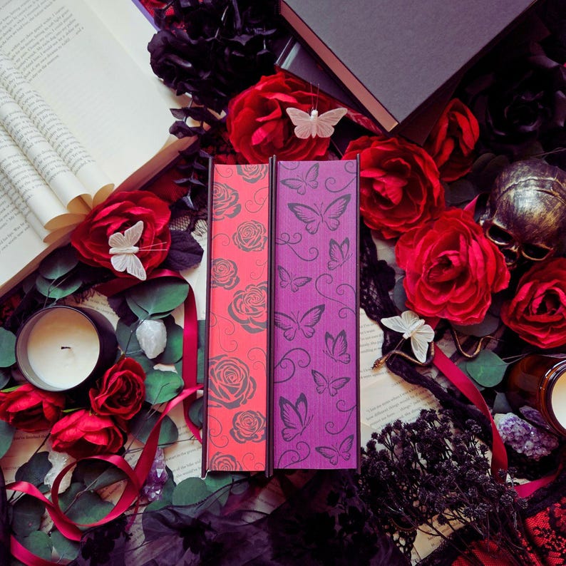 May include: Two decorative book boxes, one coral and one purple, are displayed with red roses, butterflies, and a skull. The coral box features a rose pattern, while the purple box has a butterfly design. A candle and open book are also visible.