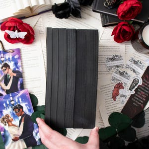 May include: A stack of black elastic bands, likely for crafting or sewing, is held over a collection of decorative items. The scene includes red roses, bookmarks, and scattered paper, creating a gothic or romantic aesthetic.