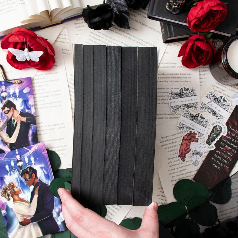 May include: A stack of black elastic bands, likely for crafting or sewing, is held over a collection of decorative items. The scene includes red roses, bookmarks, and scattered paper, creating a gothic or romantic aesthetic.