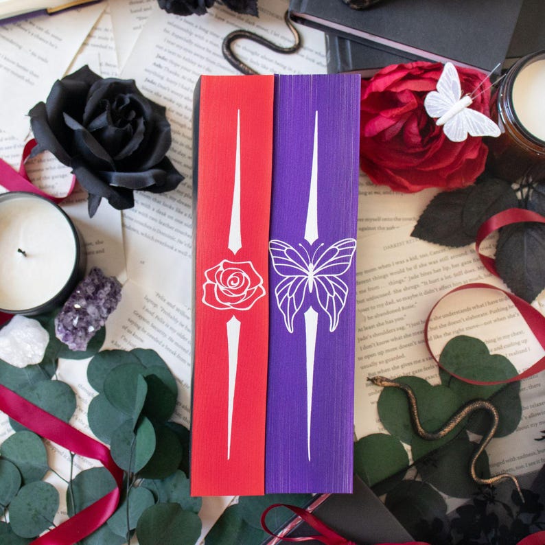 May include: Two bookmarks, one red and one purple, with white decorative designs. The red bookmark features a rose, and the purple bookmark has a butterfly. Both have a white vertical design. The bookmarks are on a background of open books and decorative items.