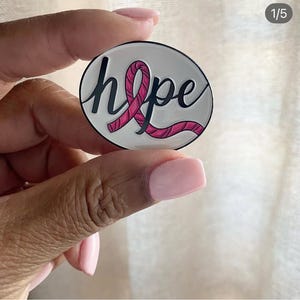 May include: A round, white enamel pin with the word "hope" in black script and a pink ribbon design. The pin is held between fingers, showcasing the design's details and colors.