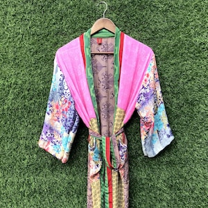 May include: A vibrant pink robe with colorful floral patterned sleeves and a patterned interior. The robe has green and red trim and a matching belt. The robe is hanging on a wooden hanger against a green background.