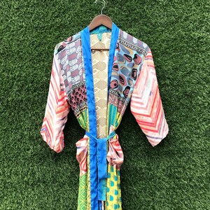 Puede incluir: Una bata de manga larga con un diseño de patchwork que presenta varios patrones coloridos, incluyendo formas geométricas, motivos florales y rayas. La prenda tiene un borde azul sólido en la abertura frontal y un cinturón a juego en la cintura.