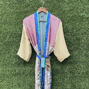 Op de afbeelding: Een kimono-badjas met lange mouwen en een patchwork-ontwerp met diverse patronen en kleuren, waaronder roze, blauw, geel en grijs. Het kledingstuk heeft een effen blauwe bies langs de opening aan de voorzijde en een bijpassende ceintuur.