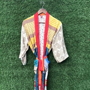 Op de afbeelding: Een kleurrijke, patroonrijke kimono met een rode kraag en ceintuur. De kimono heeft verschillende patronen, waaronder paisley, bloemen en geometrische ontwerpen, in tinten geel, bruin, blauw en oranje. Hij hangt aan een houten hanger tegen een groene achtergrond.