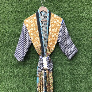 Puede incluir: Bata estampada con diseño floral en tonos amarillos, azules y blancos. Ribete negro y cinturón a juego. Mangas azul oscuro con estampado floral blanco. Colgada de una percha de madera. Diseño vibrante y colorido.