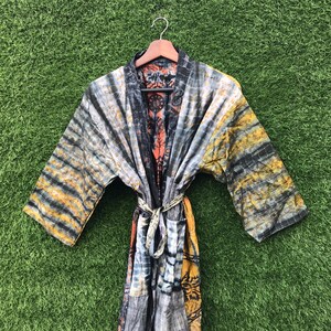 Puede incluir: Una bata con estampado tie-dye en tonos grises, negros, amarillos y naranjas cuelga de una percha de madera. La bata tiene mangas largas y un cinturón a juego. El fondo es verde.