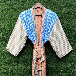 Puede incluir: Una bata estampada y colorida con cinturón. La bata presenta una mezcla de telas azules, naranjas y beige con varios diseños. Cuelga de una percha de madera sobre un fondo verde. Las mangas son anchas y el cinturón está atado.