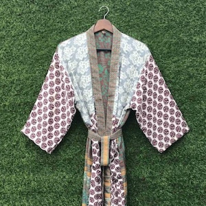 Könnte beinhalten: Ein farbenfroher Kimono-Stil-Bademantel mit floralem Muster. Der Bademantel weist eine Mischung aus Mustern und Farben auf, darunter Weiß, Burgunderrot und Grün. Er hat lange Ärmel und einen passenden Gürtel. Auf einem Holzbügel vor grünem Hintergrund.