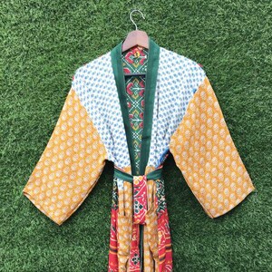 Könnte beinhalten: Ein farbenfroher Kimono-Bademantel mit grünem Kragen und Gürtel. Der Bademantel weist eine Mischung aus Mustern auf, darunter weiße Tupfen, gelbe Kreise und rot-gelbe Streifen. Der Bademantel hängt an einem Holzbügel vor grünem Hintergrund.