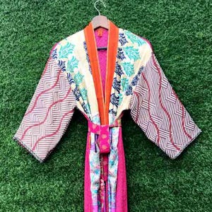 May include: A colorful, patterned kimono robe with a belt, hanging against a green background. The robe features a mix of floral and geometric designs in shades of pink, orange, cream, and blue. The sleeves have a zig-zag pattern.