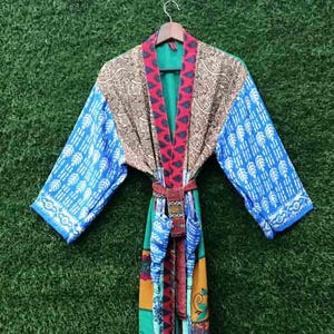 Puede incluir: Una colorida bata kimono patchwork con interior verde. La bata presenta mangas azules con un estampado de hojas blancas, un cuello estampado marrón y rojo y un cinturón a juego. La bata está colgada de una percha de madera.