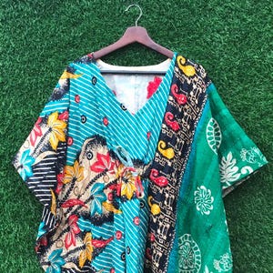 May include: A colorful, patterned kaftan dress with a V-neck and short sleeves. The dress features a mix of floral, striped, and geometric designs in teal, green, black, red, and yellow. It hangs on a wooden hanger against a green background.