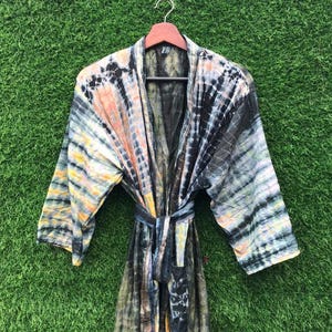 May include: A tie-dye robe with a belt, hanging on a wooden hanger. The robe features a mix of colors, including orange, black, blue, and yellow. The fabric has a striped pattern, and the robe is displayed against a green background.