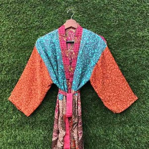 May include: A colorful, patterned robe with a floral design. The robe features a mix of orange, teal, pink, and patterned fabrics. It is hanging on a wooden hanger against a green background.