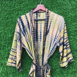 May include: A tie-dye patterned robe in shades of yellow, black, and peach hangs on a wooden hanger. The robe features a wrap-around design with a matching tie belt. The fabric has a textured appearance.