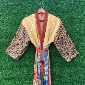 May include: A colorful, patterned kimono robe with long sleeves. The robe features a yellow floral design on the shoulders and a mix of patterns and colors throughout the garment. It has a matching belt and is hanging on a wooden hanger.