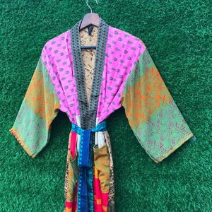 May include: A colorful patchwork robe with a floral pattern in pink, green, orange, and blue. The robe is hanging on a wooden hanger against a green background. The robe has a belt.