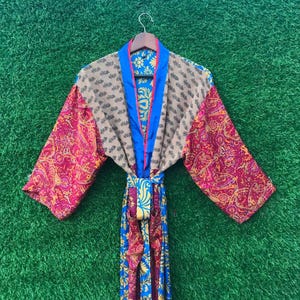 Puede incluir: Una bata colorida y estampada con un diseño de cachemira en tonos rosa, rojo y amarillo. La bata tiene un ribete azul y un cinturón a juego. Las mangas son largas y anchas, y la bata está colgada de una percha de madera.