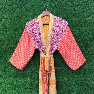May include: A colorful, patterned robe with long sleeves, hanging on a wooden hanger. The robe features a mix of orange, purple, and yellow geometric designs. A matching belt is tied at the waist. The background is green.