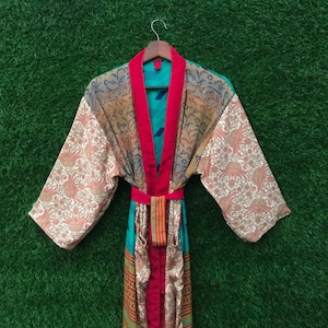 Może przedstawiać: Kolorowe, wzorzyste kimono z długimi rękawami. Szlafrok ma mieszankę tkanin w kolorze turkusowym, czerwonym i wzorzystym. Rękawy mają kremowy wzór paisley. Czerwony pasek zaciska talię. Szlafrok wisi na drewnianym wieszaku.
