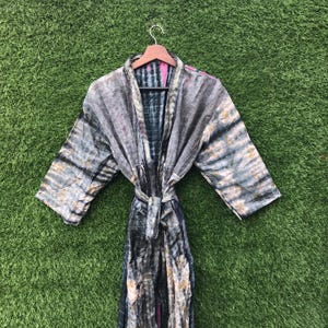 May include: A tie-dye robe in shades of gray, blue, and gold hangs on a wooden hanger. The robe features a belt and a reversible design with pink accents. The garment is displayed against a green background.