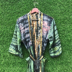 May include: A tie-dye robe in shades of green, black, and gold hangs on a wooden hanger. The robe features a belt and a patterned design. The background is a vibrant green.