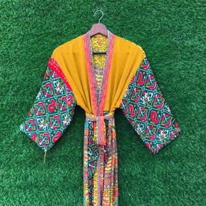 May include: A mustard yellow robe with patterned sleeves and trim, hanging on a wooden hanger. The sleeves feature a green, red, and white geometric design. The robe is tied with a matching belt, and the background is green.