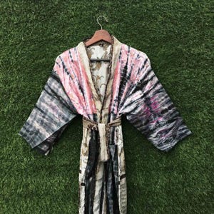 May include: A tie-dye patterned robe with pink, black, and grey stripes. The robe is hanging on a wooden hanger against a green background. The robe has a matching belt and a patterned interior.