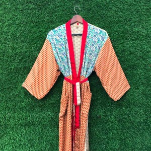 May include: A colorful robe with a red trim and belt, hanging against a green background. The robe features a mix of patterns, including orange polka dots and a blue and green floral design. The robe is made of a lightweight fabric.