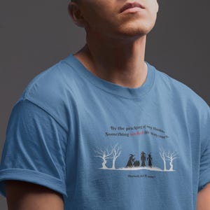 Pode incluir: Camiseta azul clara com um design gráfico. O design apresenta a citação "By the pricking of my thumbs, Something wicked this way comes." Inclui silhuetas de bruxas e árvores, com texto de Macbeth.