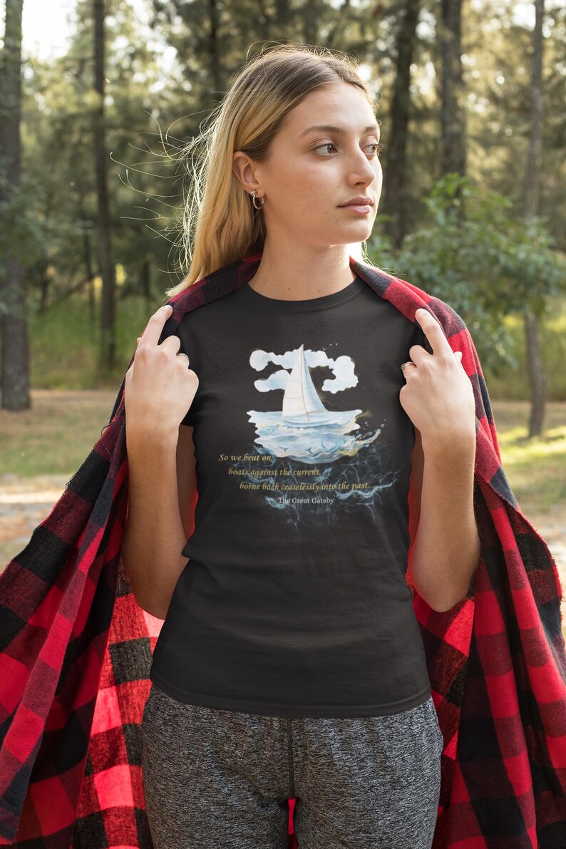 Puede incluir: Camiseta negra con un gr&aacute;fico de velero y la cita "So we beat on, boats against the current, borne back ceaselessly into the past" de El Gran Gatsby. La camiseta se usa con una franela a cuadros roja y negra.